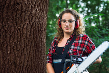 Portrait Of Lumberjack Woodcutter Standing By The Tree Trunk In The Woods Holding Chainsaw.
