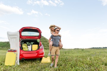 Wall Mural - Family couple having fun at the camping rolling each other on their hands running on the green grass on a summer day in the background of a car with Luggage for outdoor recreation