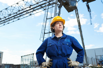 Waist up portrait of empowered woman working at construction site posing against sky with tower crane in background, copy space