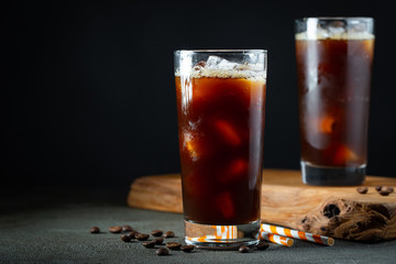 Ice coffee in a tall glass with cream poured over, ice cubes and beans on a old rustic wooden table. Cold summer drink with tubes on a black background with copy space