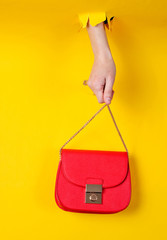 Wall Mural - Female hand is holding red leather bag over chain through torn yellow paper. Minimalistic fashion concept