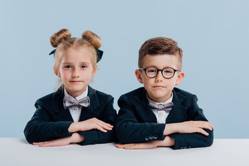 Wall Mural - education. school children look at the camera, holding hands on the table, dressed in school uniform, sitting at the white table, isolated on blue background, copy space, school concept