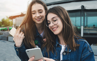 Two happy young best friends using phones on the city street