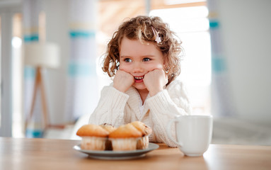 Wall Mural - A portrait of small girl with muffins sitting at the table at home.