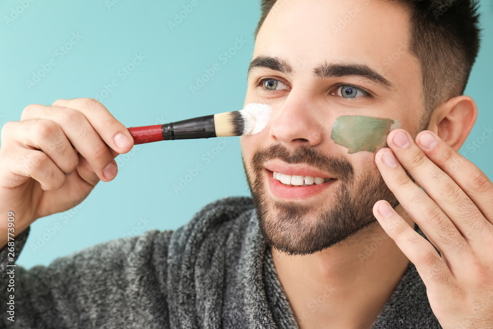 Handsome man applying clay mask onto his face against color background