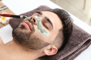 Wall Mural - Cosmetologist applying clay mask onto face of young man in beauty salon