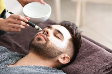 Wall Mural - Cosmetologist applying clay mask onto face of young man in beauty salon