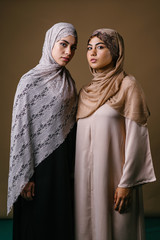 Two Muslim Middle Eastern friends in traditional dresses and hijab head scarves stand next to another to pose for a portrait together in a studio. They are both dressed conservatively but stylishly. 