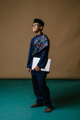 Wall Mural - Studio portrait of a confident young Asian man in traditional garb (baju melayu tunic and songkok hat) smiling as he holds his laptop computer under his arm. 