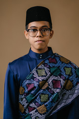 Wall Mural - Studio portrait of a happy, handsome and young Malay Asian man in a blue baju melayu hat with a patterned sash dressed for Hari Raya. He is ready to go visiting friends and family during Raya.