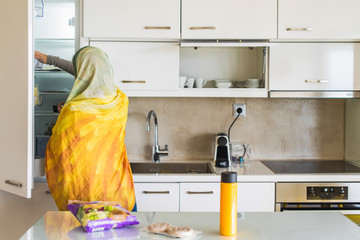 Back view on a muslim woman in a scarf taking out juice from the kitchen fridge. Breakfast preparation