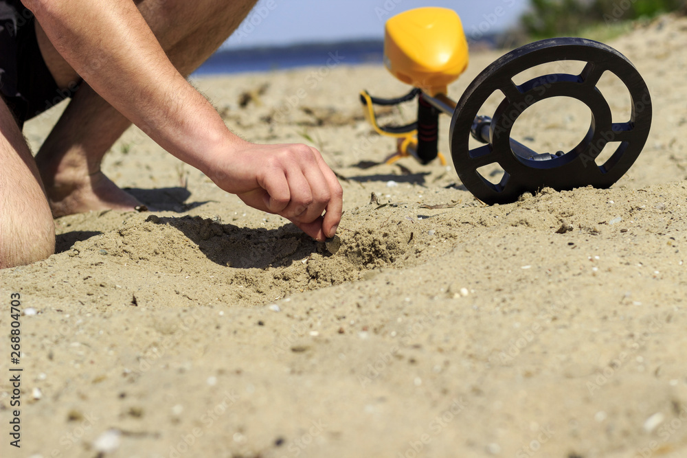A man takes a coin found in the sand with a metal detector - obrazy, fototapety, plakaty 