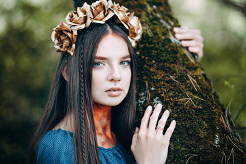 portrait of beautiful brown-haired woman in a long blue dress, with a wreath of golden roses on her head, in the fairy forest, golden paint shimmer on her neck fantasy concept