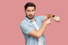 Time Is Out. Portrait Of Serious Handsome Bearded Young Man In Blue Casual Style Shirt Standing And Looking At Camera, Pointing On His Smart Watch. Indoor Studio Shot, Isolated On Pink Background.
