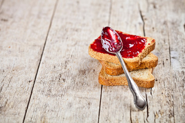 Toasted cereal bread slices stack with homemade cherry jam and spoon closeup on rustic wooden table background.