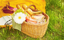 Close Up Of Picnic Basket With Food, Fruits And Flower On The Yellow Cover On The Green Grass
