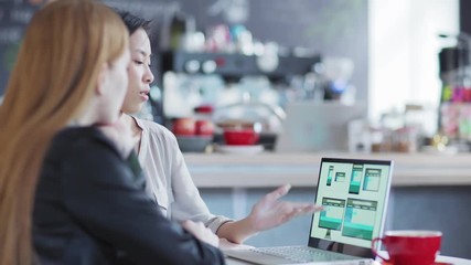 Wall Mural - Zoom out of two young businesswomen discussing website templates presented on laptop screen sitting at table in cafe, Asian woman explaining design to Caucasian colleague