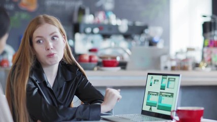 Wall Mural - Zoom out of two young businesswomen discussing website templates for different devices presented on laptop screen sitting at table in cafe, focus on woman explaining design to Asian colleague