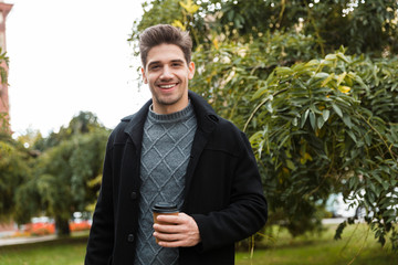 Poster - Photo of handsome man holding paper cup of takeaway coffee while walking through green park