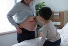 Little Asian Boy Make Painting On His Mother's Pregnant Belly In Bedroom.