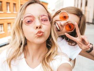 Two young smiling hipster blond women in summer clothes. Girls taking selfie self portrait photos on smartphone.Models posing on street.Female making duck face and shows peace sign