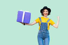 Happy Attractive Young Girl In Hipster Wear In Denim Overalls And Black Hat Standing And Holding Big Heavy Gift Box With Toothy Smile, Show Strong Arm, Looking At Camera. Studio Shot, Green Background
