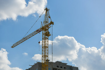 high-rise construction crane with a long arrow of yellow color against the blue sky over a new multi-storey building of concrete and brick under construction