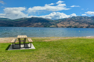 Wall Mural - Picnic table with benches on Okanagan lake shore. Table and benches at rest area close to waterfront with mountains view on cloudy and blue sky background.