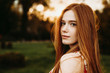 Portrait of a beautiful redhead woman with green eyes and freckles looking at camera against sunset outside over the shoulder.