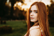 Portrait Of A Beautiful Redhead Woman With Green Eyes And Freckles Looking At Camera Against Sunset Outside Over The Shoulder.