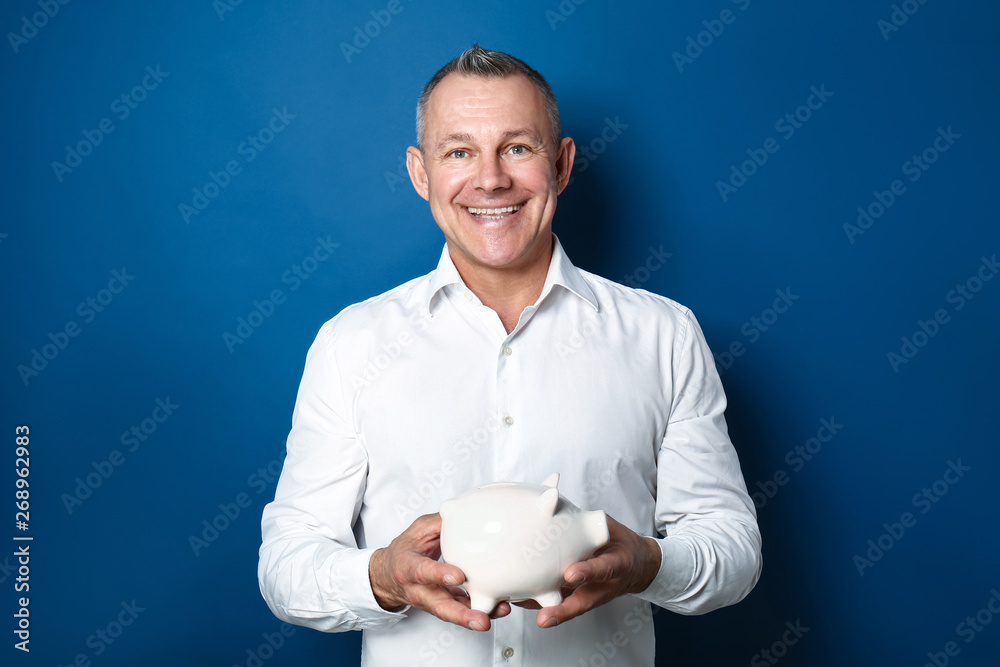 Handsome middle-aged man with piggy bank on color background