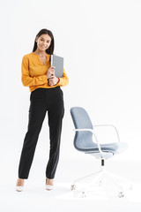 Sticker - Full length portrait of charming young businesswoman dressed in elegant wear holding paper folder and standing by chair in office