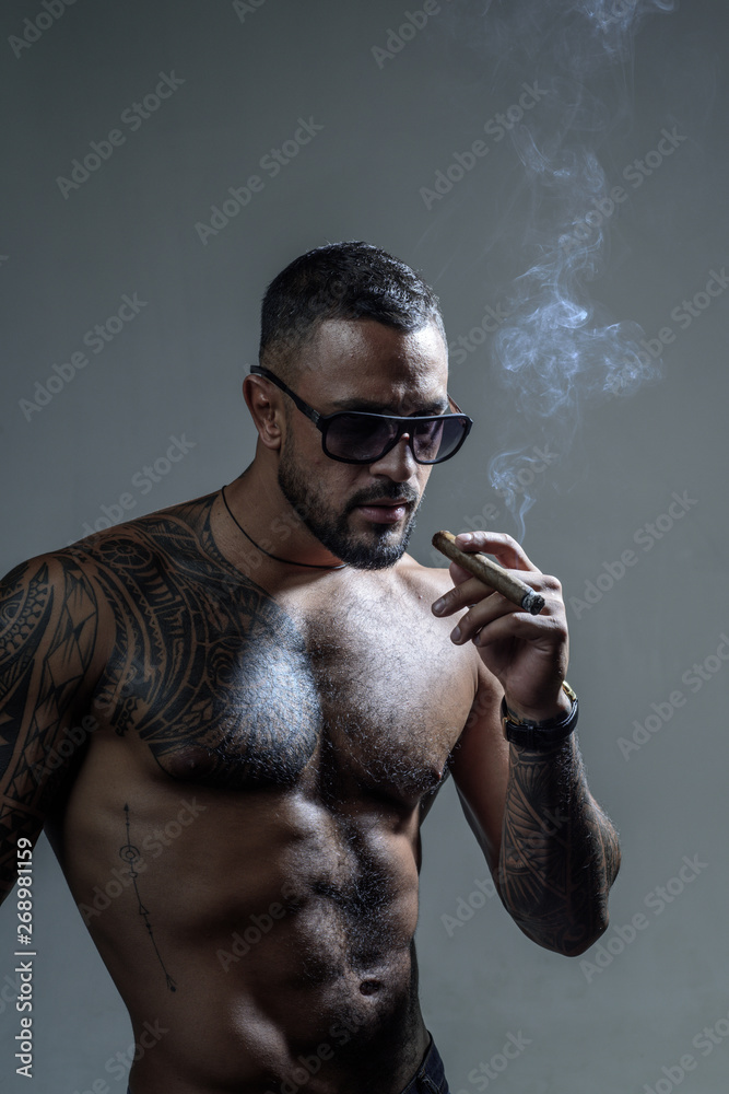 Expensive tobacco. Cigar smoking enjoy life moment. Bearded muscular macho  beautiful torso fashion sunglasses smoking cigar. Elite tobacco. Pure  enjoyment. Consuming tobacco. Tattooed man with cigar Stock Photo