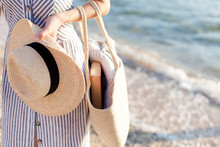 Woman At Sea Beach. Girl Holds Straw Hat And Bag With Book And Plaid. Traveler Is Enjoying Summer Vacation And Holidays.