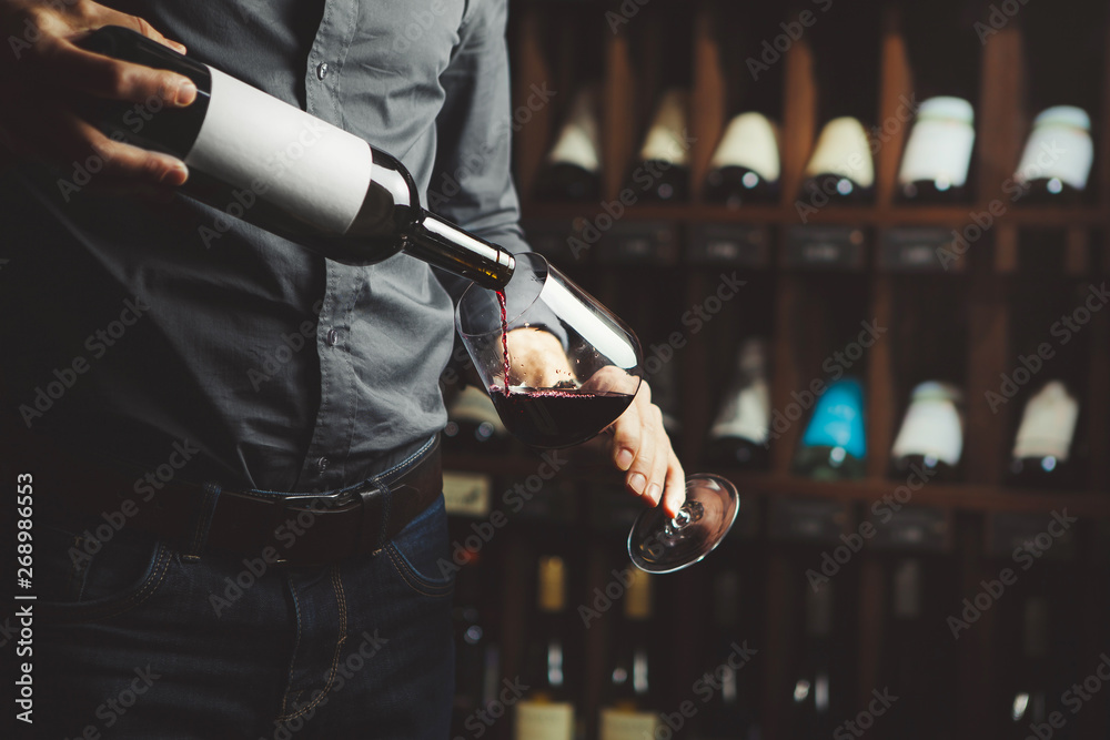 Close up shot of sommelier pouring red wine from bottle in glass on underground cellar background - obrazy, fototapety, plakaty 