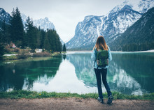 Young Woman With Backpack Is Standing On The Coast Of Mountain Lake At Cloudy Day In Spring. Travel In Italy. Landscape With Slim Girl, Reflection In Water, Snowy Rocks, Green Trees. Vintage Toning