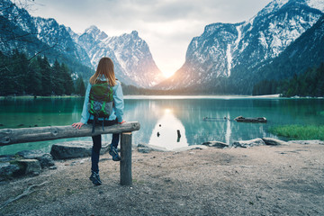 Young woman with backpack is sitting on the coast of mountain lake at sunset in spring. Travel in Italy. Landscape with slim girl, reflection in water, snowy rocks, green trees. Vintage toning