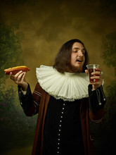 Young Man As A Medieval Knight On Dark Studio Background. Portrait Of Male Model In Retro Costume. Holding A Glass Of Beer And Hot-dog. Human Emotions, Comparison Of Eras, Facial Expressions Concept.