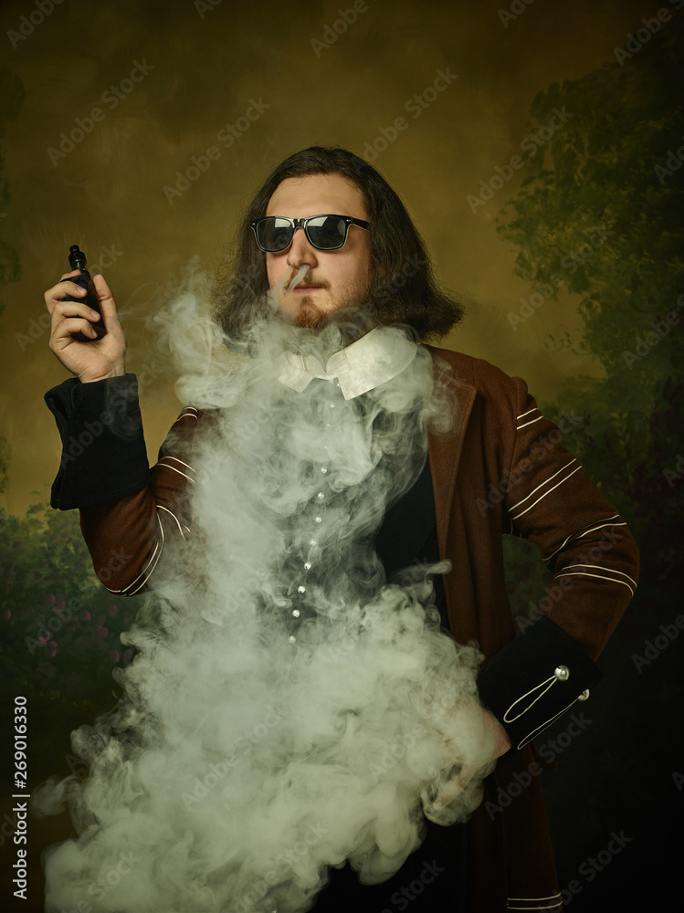 Foto Young man as a medieval knight in sunglasses on dark studio  background. Portrait of male model in retro costume. Smoking vape. Human  emotions, comparison of eras and facial expressions concept. do