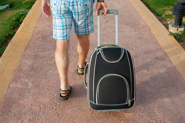  Young man pulling suitcase on road in summer park. Travelling guy wearing smart shorts walking away with his luggage