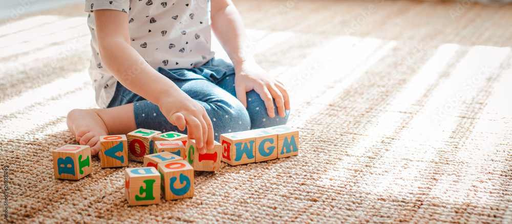 child plays with wooden blocks with letters on the floor in the room a little girl is building a tower at home or in the kindergarten. - obrazy, fototapety, plakaty 