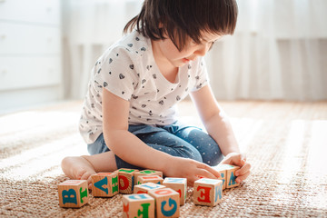 Wall Mural - child plays with wooden blocks with letters on the floor in the room a little girl is building a tower at home or in the kindergarten.