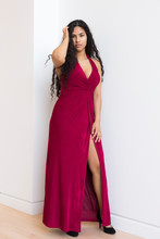 Vertical Full-length Portrait Of Gorgeous Sultry Hispanic Young Woman Wearing Elegant Evening Red Dress With High Slit And Touching Her Long Hair While Leaning Against White Wall