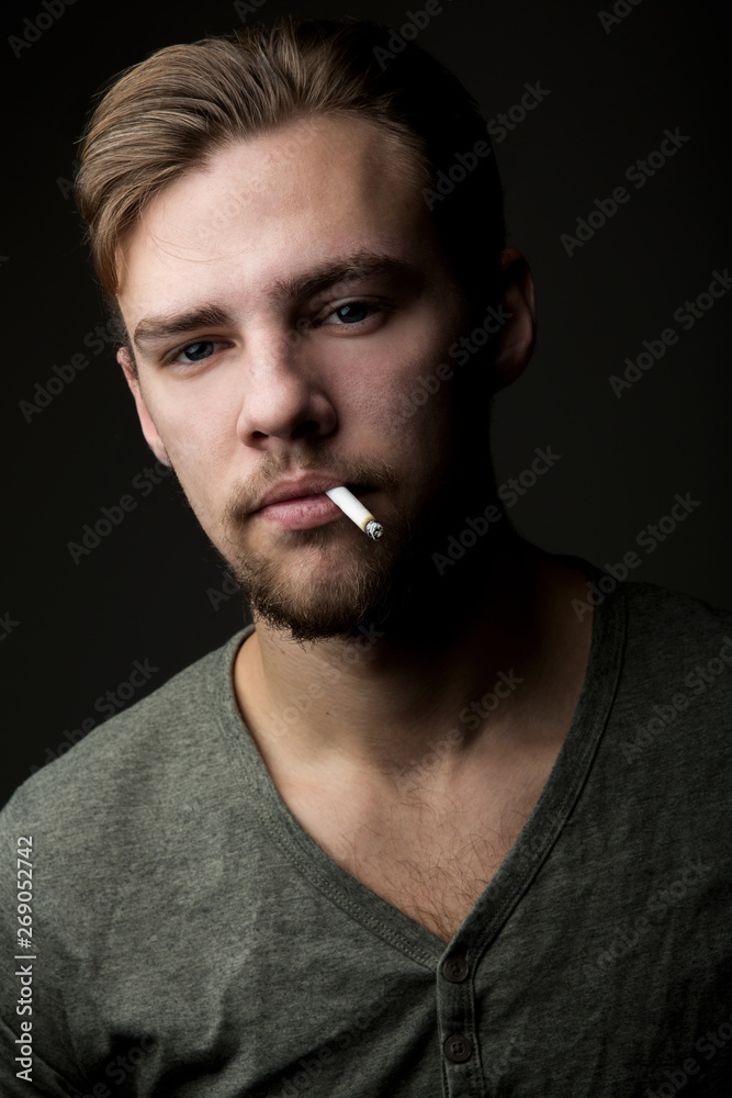 Portrait of a young Caucasian Smoking man with a cigarette in his mouth and  short beard and mustache on a dark background Stock Photo