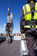 Professional industrial climber in helmet and uniform holds telecomunication equipment in his hand and antennas of GSM DCS UMTS LTE bands, outdoor radio units, optic fibers, power cables are installed