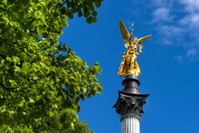 Germany, Munich, Bogenhausen: Steeple Of The Peace Column With Famous Golden Angel Of Peace Statue (Friedensengel) At The Top In The City Center Of The Bavarian Capital With Green Trees And Blue Sky.
