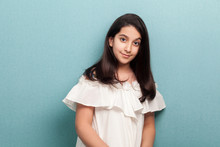 Portrait Of Happy Calm Beautiful Brunette Young Girl With Black Long Straight Hair In White Dress Standing And Looking At Camera With Smile. Indoor Studio Shot Isolated On Blue Background.