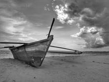 A Small Black And White Photography Of A Traditional Fishing Canoe On The Beach During The Low Tide.