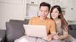 © tirachard - Asian couple using laptop for search web in living room at home, sweet couple enjoy love moment while lying on the sofa when relax at home. Lifestyle couple relax at home concept.