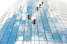 Five Window Washers Work At A Height On A High-rise Building With A Glazed Facade Against A Blue Sky With Light Clouds.